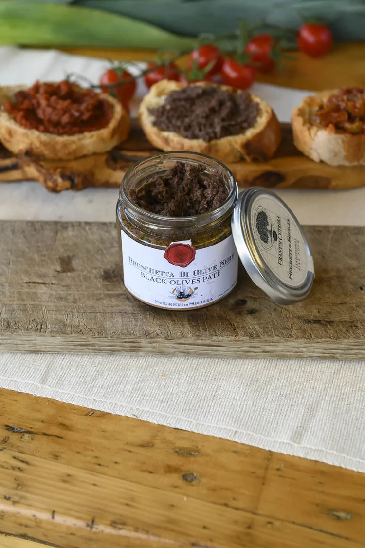 Jar of black olive paste on a wooden board with bread and tomatoes in the background